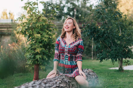 Young adult woman, with eyes closed, sitting on a tree in the forest, enjoying the tranquility of relaxed nature. concept of clean air, freedom and environmental activism.の写真素材