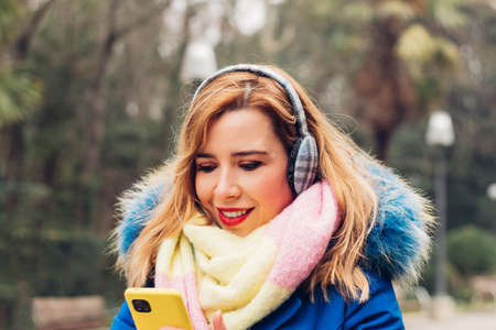 Portrait of a charming smiling young adult woman typing a message on the mobile phone on a winter day wearing lots of warm clothes, colorful.の写真素材