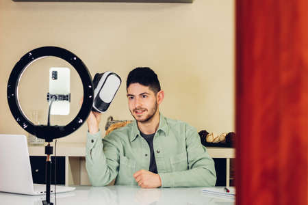 Young adult man showing off a virtual reality goggles behind the scenes. Handsome man recording a tutorial giving his opinion about a product. Camera setup and lighting.の写真素材