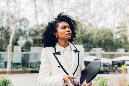 Front view of a businesswoman in white modern suit with curly hair holding her folder by looking up...