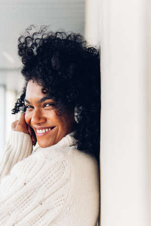 Vertical image of a cheerful young woman with afro hair in white sweater leaning on wall looking at camera.の写真素材