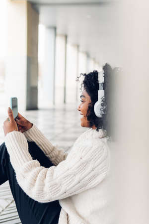 Vertical image of cheerful young woman sitting on floor looking at her phone and listening to music with her wireless headphones.の写真素材
