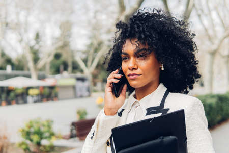 Portrait of a businesswoman holding work folders while talking on phone outdoors.の写真素材