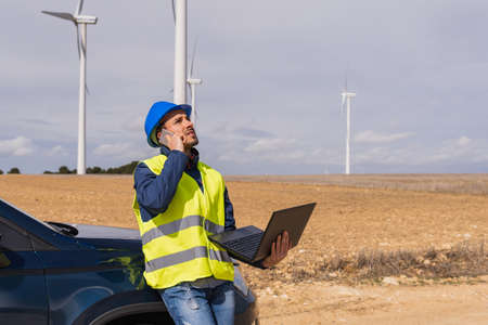 Engineer in work clothes, on his company car, talking on his mobile phone and a laptop in hand at...