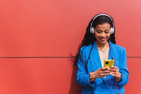 Young african woman wearing blue clothes on red background, using her mobile phone, listening to music with wireless headphones. Copy space.の写真素材