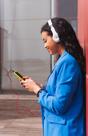 Smiling African girl using her mobile phone outdoors.Vertical shot of a 20 year old girl listening to music leaning against a red wall.の写真素材