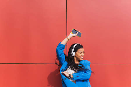 African ethnicity student happy and smiling while dancing and listening to music with energy and joy, celebrating weekend, vacation or school grades. Blue clothes and red color background wall.の写真素材
