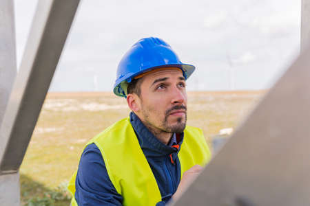 Concentrated bearded male engineer in safety vest and hardhat looking up while standing near metal railing on field during work in countrysideの写真素材