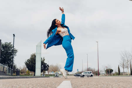 Full body side view of positive African American female looking up while standing on tiptoes on asphalt roadway in cityの写真素材