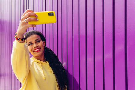 Happy girl taking a selfie portrait with her yellow smartphone on a purple background. Young African...