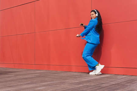 Full body side view of positive African American female in blue suit dancing while listening to music in headphones on red backgroundの写真素材