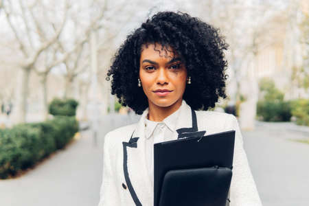 Charming African American businesswoman with folder in hands in formal wear looking at camera while standing on street of cityの写真素材