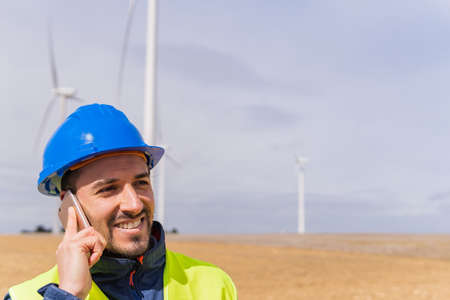 Happy man in blue hardhat smiling and looking away while answering phone call against windmills and...