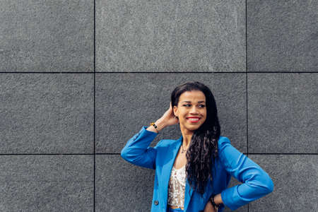 Happy young ethnic female in casual outfit smiling and looking at camera while standing against gray wall on city streetの写真素材