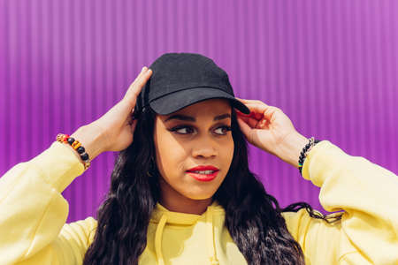 Stylish young female in trendy outfit touching head and looking at camera while standing against violet backgroundの写真素材