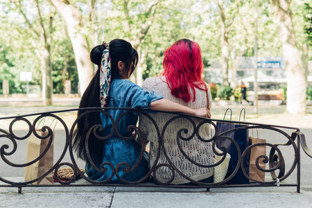 Back view of anonymous young women in casual clothes sitting on bench between shopping bags and embracing each other on sunny weekend day in parkの写真素材