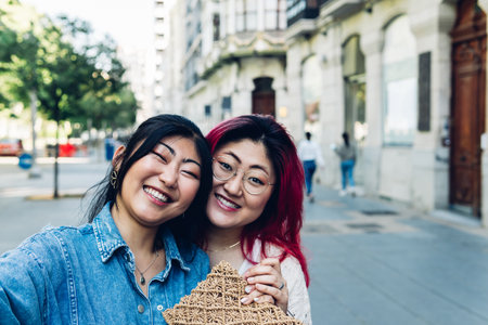 Happy young Asian women in casual clothes smiling and looking at camera while taking selfie on blurred background of city street on sunny summer dayの写真素材