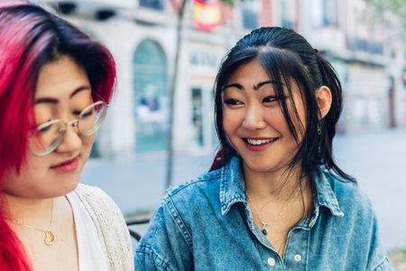 Cheerful young Asian female in denim shirt smiling and supporting sad sister while resting on bench on weekend day on city streetの写真素材