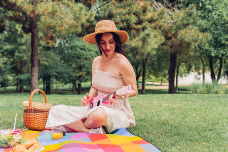 Mid adult brunette woman playing a pink ukulele at a picnic with friends. Young adult girl playing music in the countryside in a fun and care free way.の写真素材