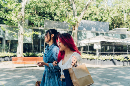 Young Asian woman in casual clothes with paper bags holding arm of best friend and walking on city street after shopping on sunny summer dayの写真素材