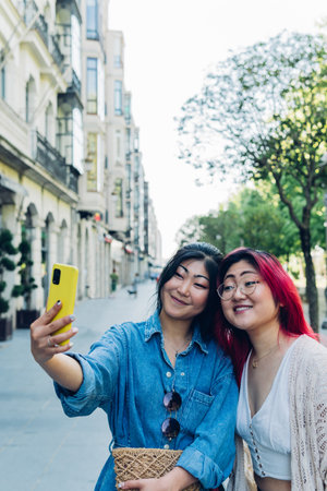 Happy young Asian girlfriends in casual clothes smiling and taking selfie via smartphone while standing on city street on sunny weekend day togetherの写真素材