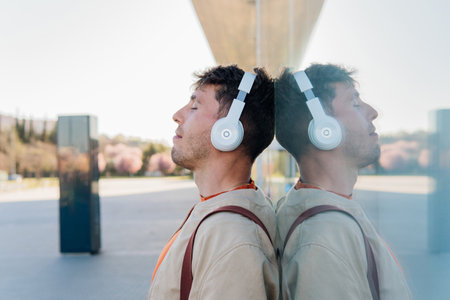 Side view of young male reflecting in glass wall while listening to songs on headphones against blurred city on sunny dayの写真素材