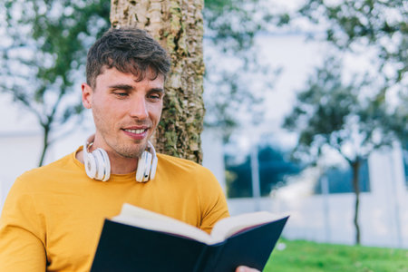 Positive young male in casual clothes smiling and reading book while sitting near tree on sunny day in parkの写真素材