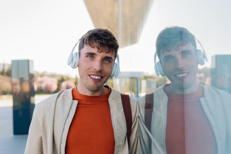 Positive young male in casual clothes and headphones smiling and looking at camera while standing on city streetの写真素材
