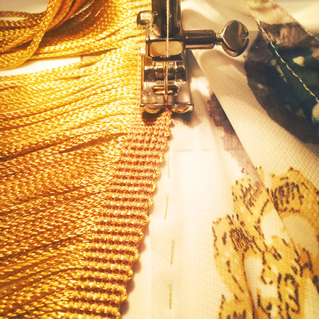 The image shows the foot of a sewing machine that is joining a patterned silk to a strip of gold fringe. The lighting is typical of the sewing machine.の写真素材