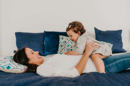 young pretty mother plays with her daughter in bed at homeの写真素材