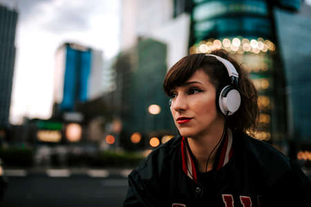 Beautiful spanish woman listening to music with earphones and smart phone outdoors. Latin girl in sport clothes with curly hairstyle in urban background. night cityの写真素材