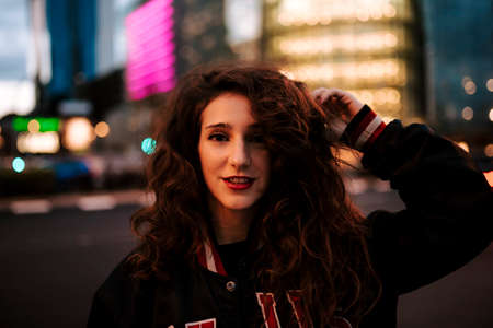 Beautiful spanish woman listening to music with earphones and smart phone outdoors. Latin girl in sport clothes with curly hairstyle in urban background. night cityの写真素材