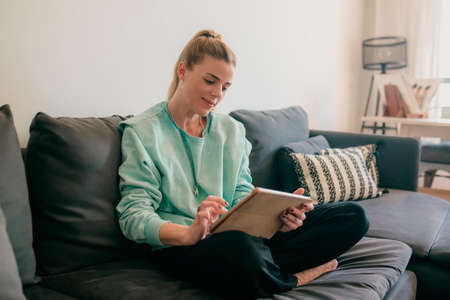 Happy beautiful woman watching videos or enjoying entertainment content on a tablet sitting on the...