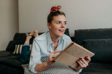 Happy beautiful woman watching videos or enjoying entertainment content on a tablet sitting on the sofa in the house.の写真素材
