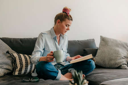 Beautiful smiling woman reading a book and lying on the sofa in the living room. Have a good time at home.の写真素材