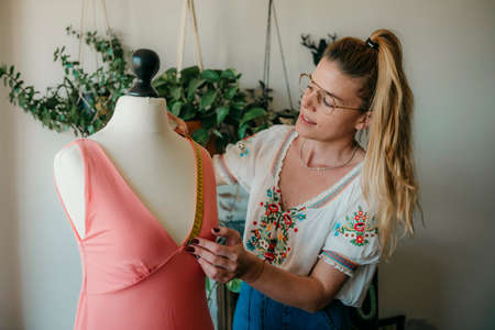 Young woman fashion designer measuring mannequin, seamstress holding tape working with dummy in cozy home or tailor shop, dressmaking and sewing concept, exclusive unique clothes creationの写真素材