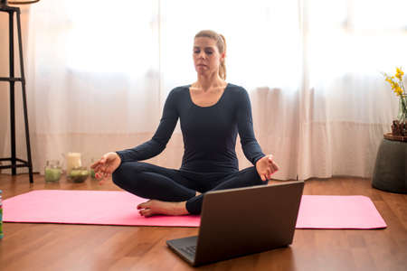 Beautiful blonde woman doing home workout indoors. Woman practice yoga at home. Fit girl using workout tutorials for healthy active lifestyle. Woman using quarantine for home workouts. Stretching in homeの写真素材