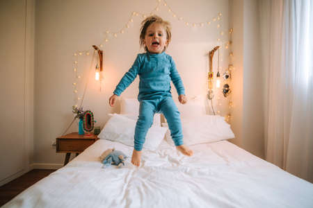 little blond boy has fun jumping and playing on a big bed. family at home. coronavirus. quarantine.の写真素材