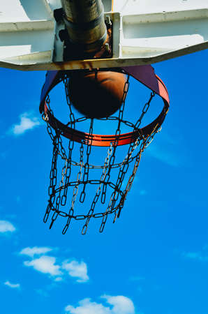 A basketball hoop with a basketball. The basketball is going in the hoop. The hoop has a double rim and a chain net. It's shot from below with the sky as a background.の写真素材