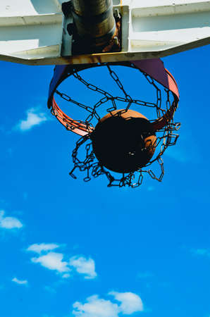 A basketball hoop with a basketball. The basketball is going in the hoop. The hoop has a double rim and a chain net. It's shot from below with the sky as a background.の写真素材
