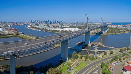 A high-angle aerial view of the West Gate Bridge spanning the Yarra River in Melbourne, Australia. The image captures busy highway traffic, the industrial port district, and the distant CBD city skyline under a clear blue sky.の写真素材
