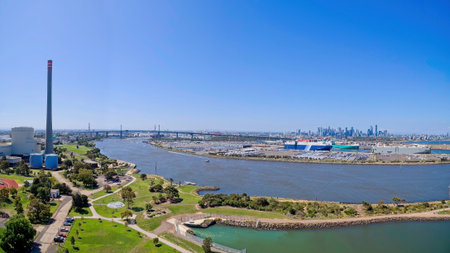 A wide aerial panoramic view of the entrance to the Port of Melbourne. The scene features the Yarra River, industrial shipping docks with large cargo vessels, the Newport Power Station chimney, and the West Gate Bridge leading to the distant city skyline.の写真素材