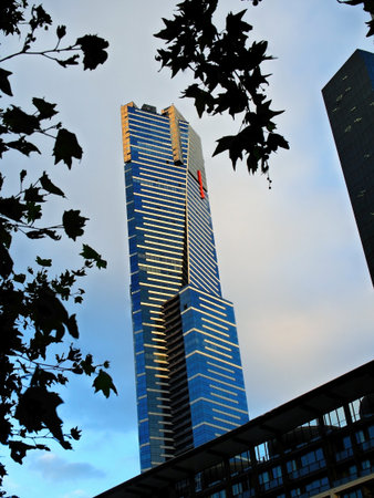 A low-angle view of a modern supertall residential skyscraper (Eureka Tower) in Melbourne, featuring its distinctive blue glass, framed by silhouetted tree leaves against the sky.の写真素材