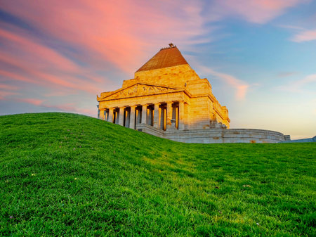 A low-angle view of the Shrine of Remembrance in Melbourne, Australia, glowing in the warm light of sunset. The historic war memorial sits atop a grassy hill against a dramatic pink and blue sky.の写真素材