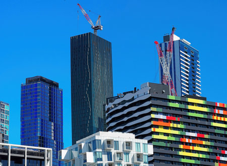A vibrant view of modern urban architecture featuring a distinctive colorful residential building with red, yellow, and green panels, contrasted against blue glass skyscrapers and construction cranes under a blue sky.の写真素材