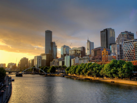 A scenic sunset view of the Melbourne city skyline, featuring the historic Flinders Street Station, the Yarra River, and the pedestrian footbridge bathed in golden light.の写真素材