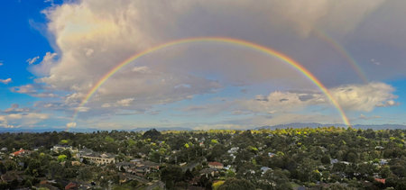 A spectacular panoramic view of a complete double rainbow stretching across the sky over a leafy residential suburb in Melbourne, Australia. The scene captures the colorful spectrum against dramatic storm clouds and green trees.の写真素材