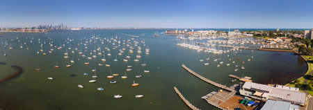 A wide aerial panoramic view of the Williamstown harbor filled with hundreds of moored sailboats and yachts. The Melbourne CBD city skyline and industrial port cranes are visible in the distance across Hobsons Bay on a sunny day.の写真素材