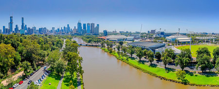 A wide aerial photograph capturing the Melbourne city CBD skyline in the background, with the Yarra River, rowing crews, parklands, and major sporting stadiums in the midground under a sunny blue sky.の写真素材