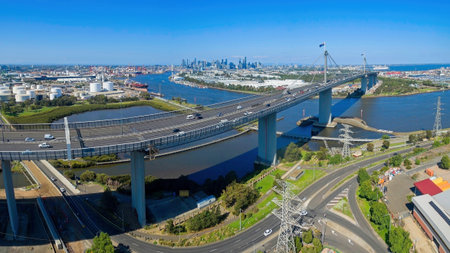 A high-angle aerial view of the West Gate Bridge spanning the Yarra River in Melbourne, Australia. The image captures busy highway traffic, the industrial port district, and the distant CBD city skyline under a clear blue sky.の写真素材
